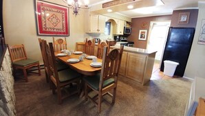 A dining area with a wooden table set for six, surrounded by wooden chairs with green cushions. A chandelier hangs above the table, and a framed quilt is displayed on the wall.