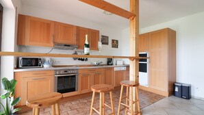 A modern kitchen with wooden cabinets, a microwave, stove, and a refrigerator. There are two wooden stools and a potted plant. - Large, bright apartment right next to the forest with a beautiful view (Welschneudorf)