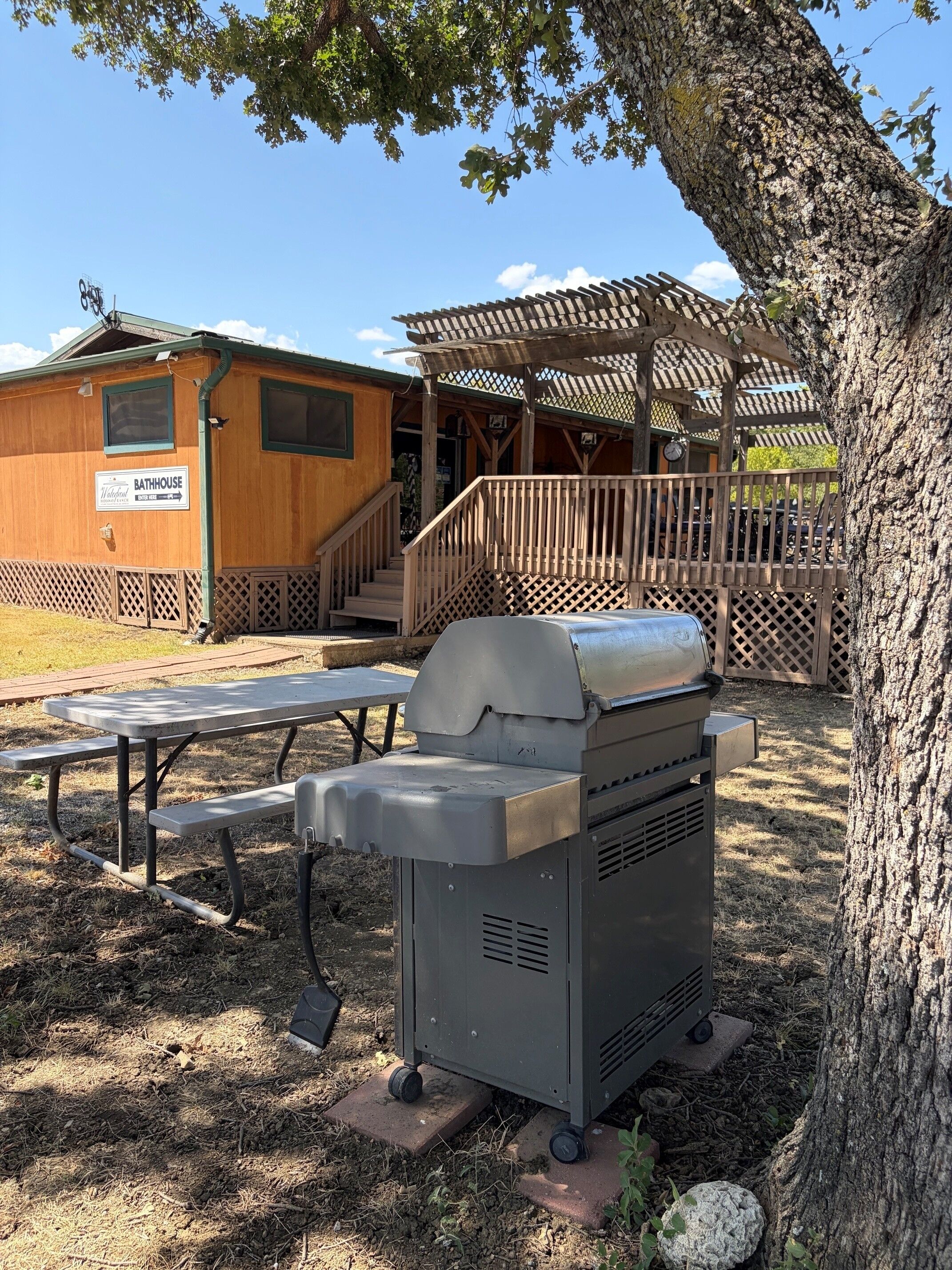A portable grill and picnic table in a campsite setting with a wooden building labeled 'Bathhouse' in the background.