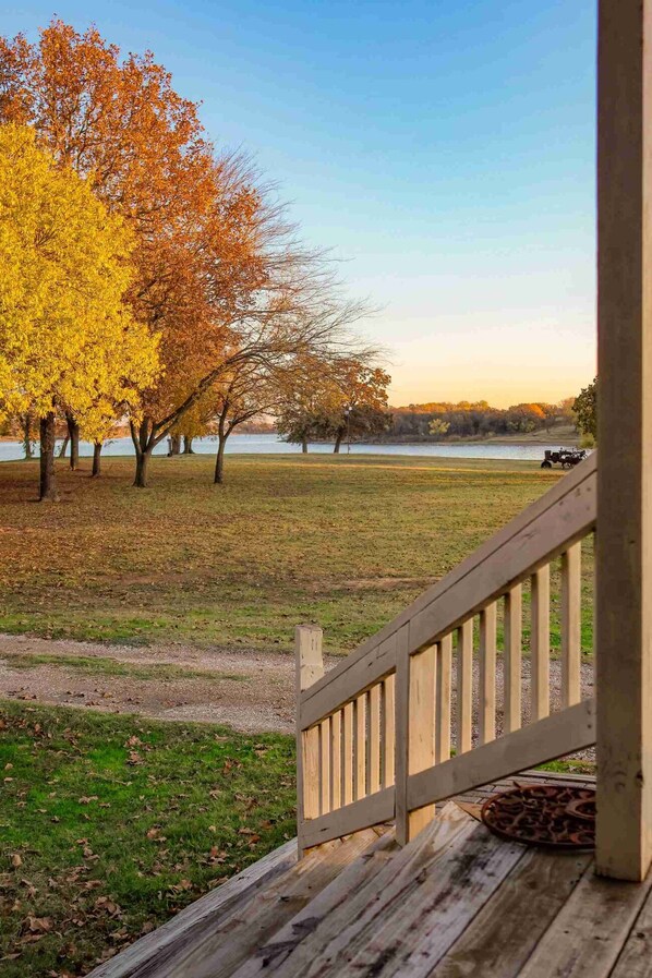 A wooden deck with a railing, a grassy field, and trees with autumn foliage. - Lakefront Escape on 14+ Private Acres | Fish, Swim & Relax (Little Elm)