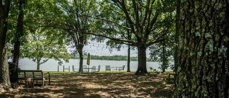 A park with a bench, tables, and chairs under trees by a lake.