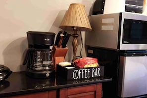 A coffee station with a coffee maker, a coffee bar sign, a bag of coffee, and a lamp.