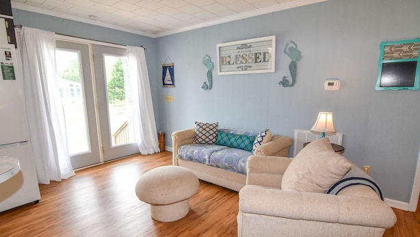 A living room with a sofa, ottoman, and a lamp. There is a window with curtains, a wall clock, and a framed sign.