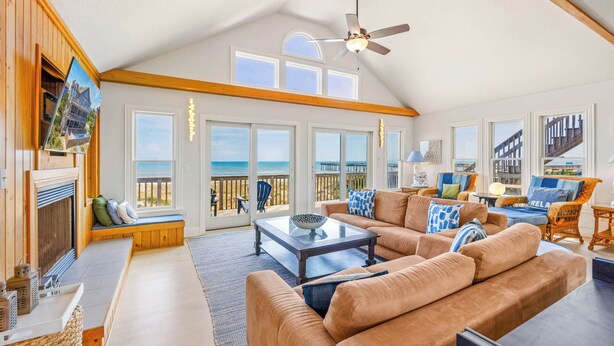 A spacious living room with a large window overlooking the beach, a ceiling fan, and a cozy seating area with a fireplace.