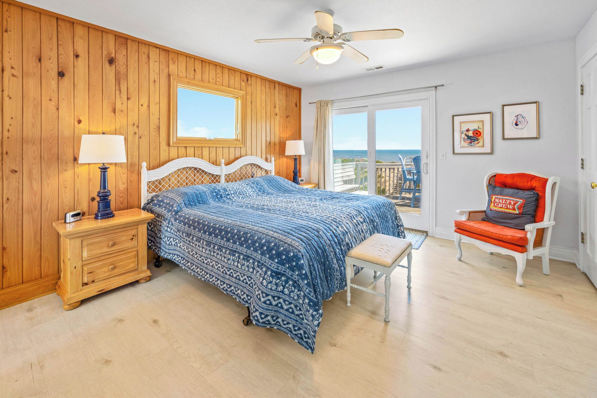 A bedroom with a wooden paneled wall, a bed with a patterned blue bedspread, a nightstand with a lamp, a chair with a cushion, and a balcony view.