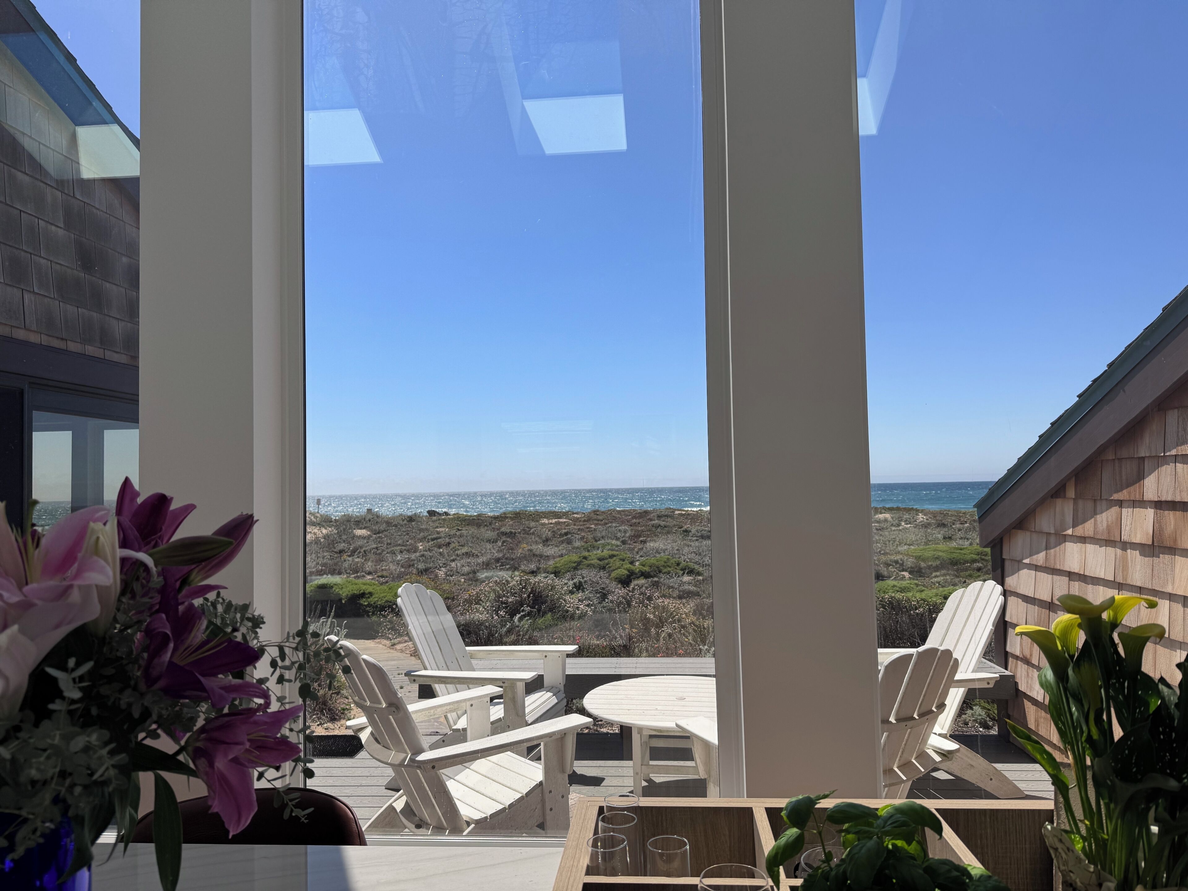 A patio with Adirondack chairs, a table, and a view of the ocean.