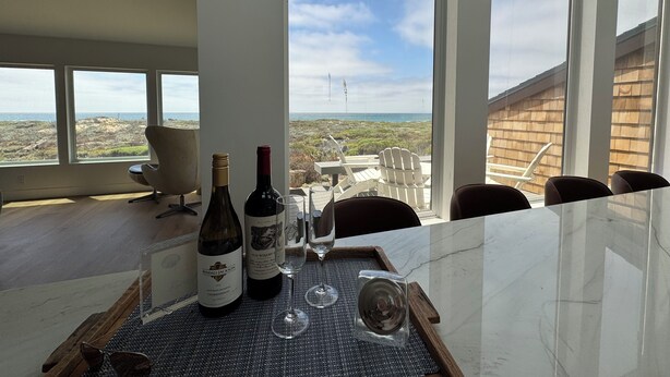 Un comedor con una mesa preparada para dos personas, que incluye una botella de vino, una copa y una bandeja. Amplias ventanas ofrecen vistas al paisaje y a una estructura de madera.
