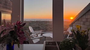 Una habitación con vista al atardecer sobre el océano, una mesa de comedor con sillas y un jarrón de flores.