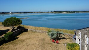 A coastal view with a clear sky, a sandy area with a small tree, and a building with a stone facade.