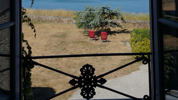 A view through a glass door of a lake with chairs and a small tree in the foreground.