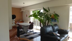 Een woonkamer met een zwarte leren bank, een eettafel met stoelen, een grote kamerplant en uitzicht door een glazen deur.