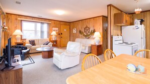 A cozy living room with a sofa, two white armchairs, a coffee table, a TV, and a dining table with chairs.