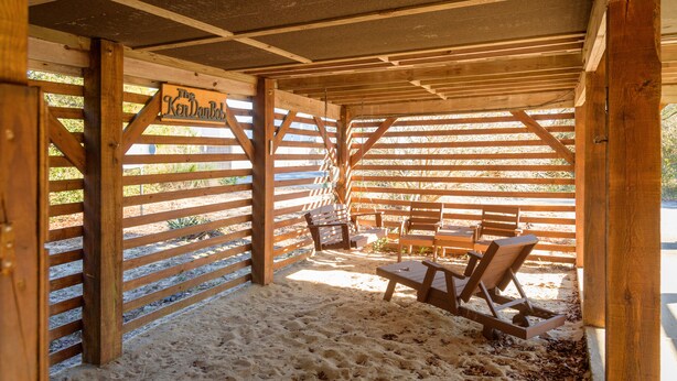 A wooden beach cabana with a sign reading "The Ken DanBob." It features a sandy floor, wooden benches, and a swing.