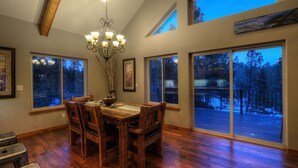 A dining area with a wooden table and chairs, large windows with a view of a snowy deck, and a chandelier. - Shiprock Cabin -  Modern 3 Bedroom 3.5 bathroom cabin with fire table (Nemo)