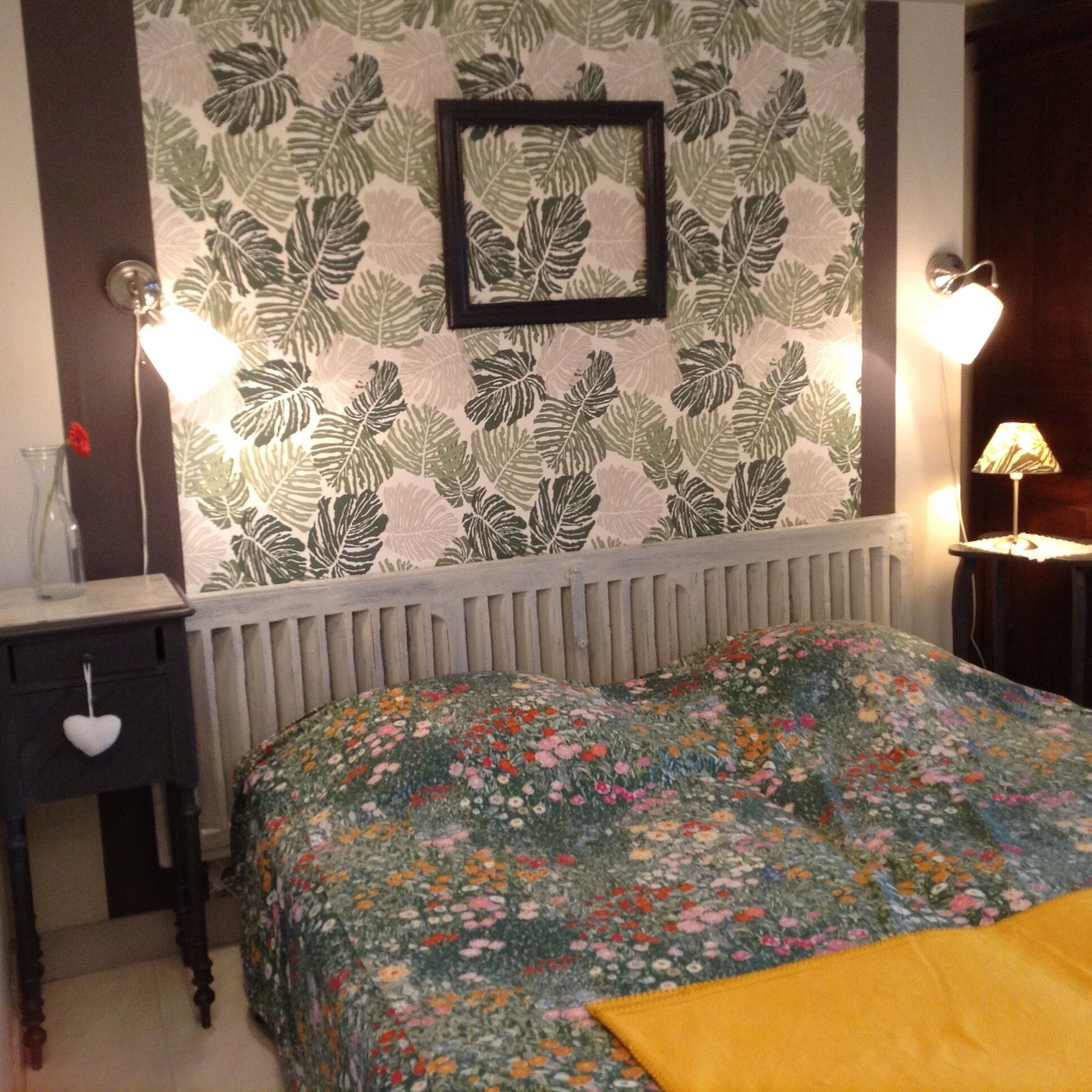 A bedroom with a floral-patterned wall, a framed picture, a bedside table with a lamp, and a bed with a floral bedspread.