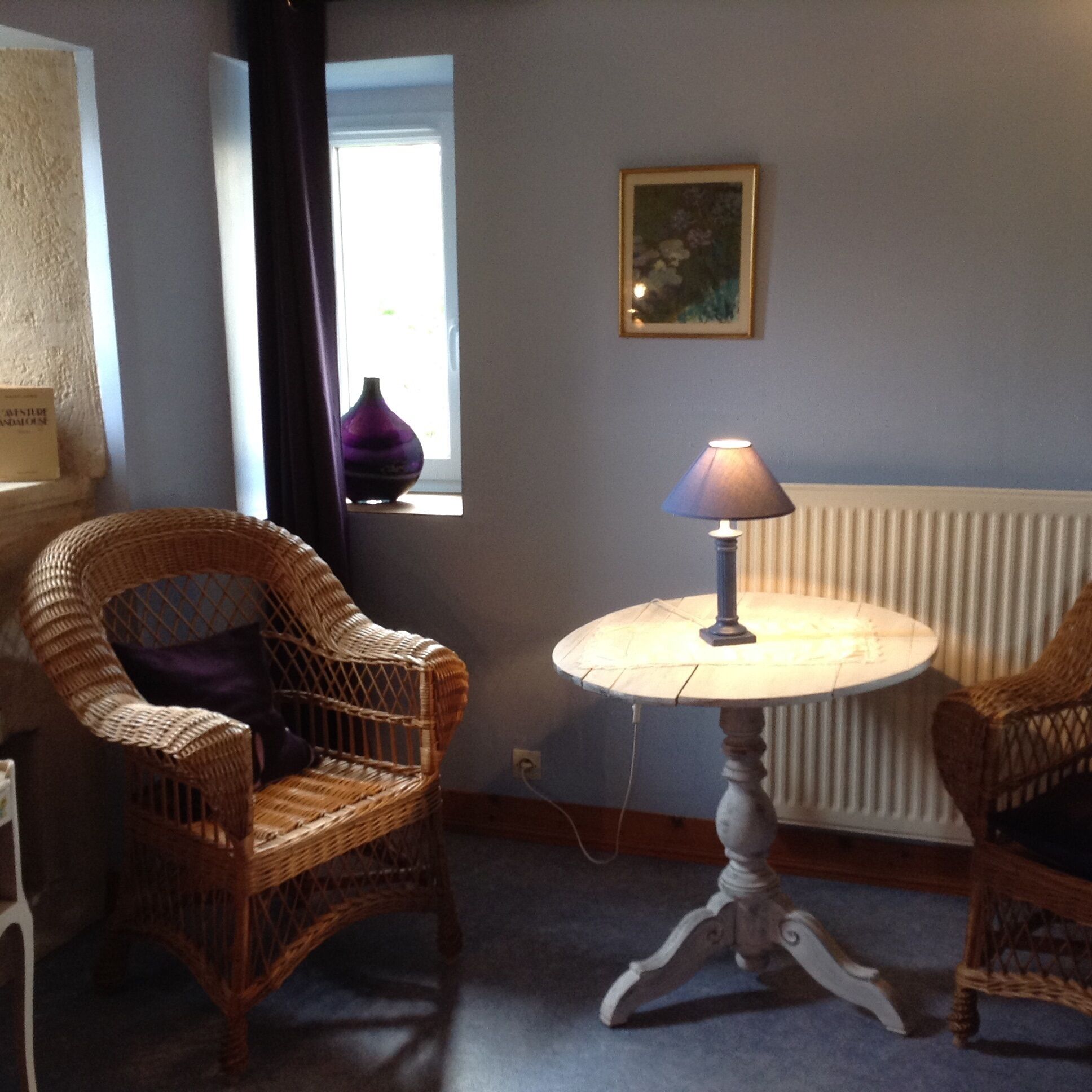 A room with wicker chairs, a table with a lamp, a window with a vase, and a radiator.
