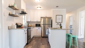 A modern kitchen with white cabinetry, a stainless steel refrigerator, a microwave, and a sink. There are wooden shelves above the countertop and a green stool in the corner. - Bright, Spacious, Family-Friendly, 8 Blocks to Main St (Fredericksburg)