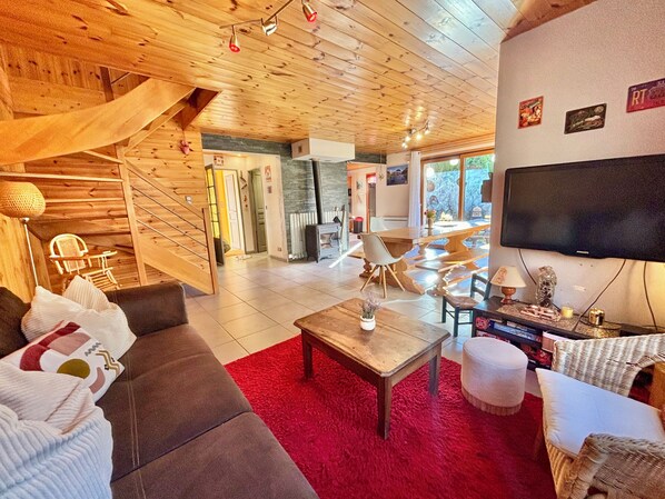 A spacious living room with a wooden ceiling, a large flat-screen TV, a red rug, and a staircase leading to another level.