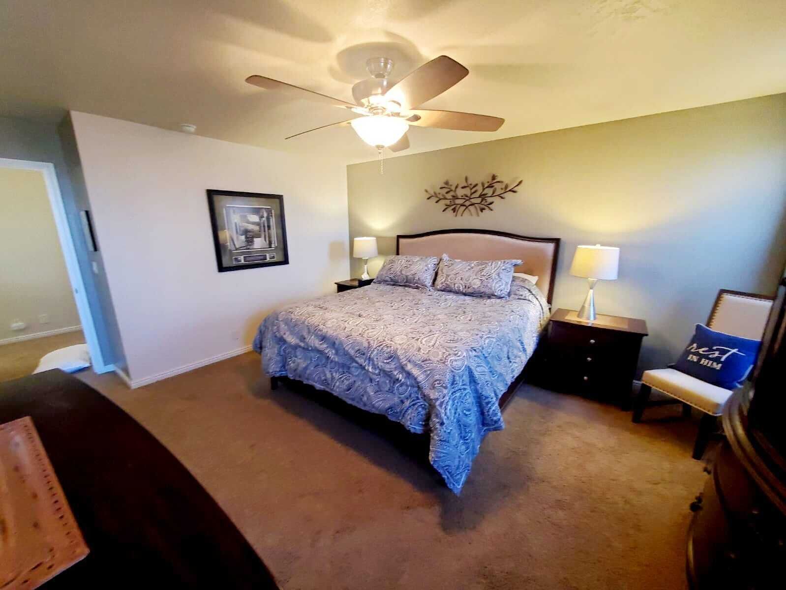 A bedroom with a bed, nightstands, a ceiling fan, a wall-mounted artwork, and a chair with a cushion.
