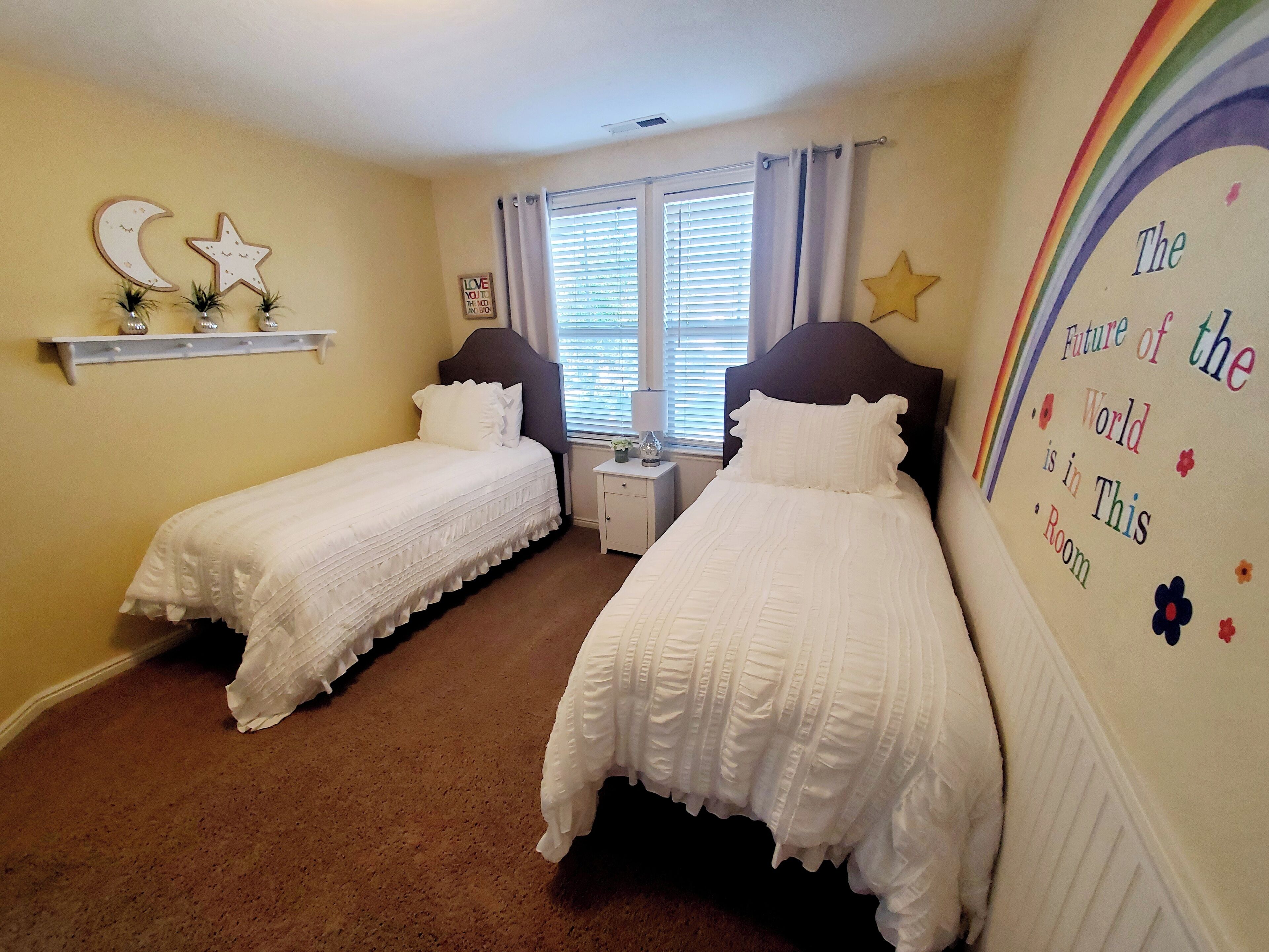 A bedroom with two single beds, a window with blinds, a nightstand with a lamp, and a wall decoration.