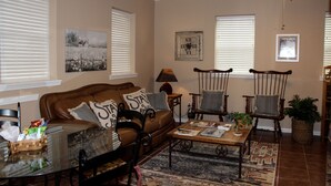 Ein Wohnzimmer mit einem braunen Ledersofa, zwei Holzstühlen, einem Couchtisch und einem Esstisch. Zwei Fenster mit Jalousien, eine Wanduhr und ein Deckenventilator sind ebenfalls vorhanden.