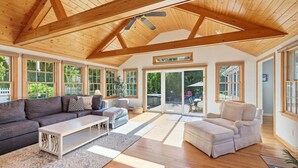 Ein geräumiges Wohnzimmer mit hoher Decke, Holzbalken, großen Fenstern und einer Schiebetür, die zu einem Außenbereich führt.