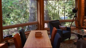 A room with large windows offering a forest view, a wooden table, two chairs, and a tripod with a camera. - Romantic island retreat  surrounded by majestic trees (Shelton)