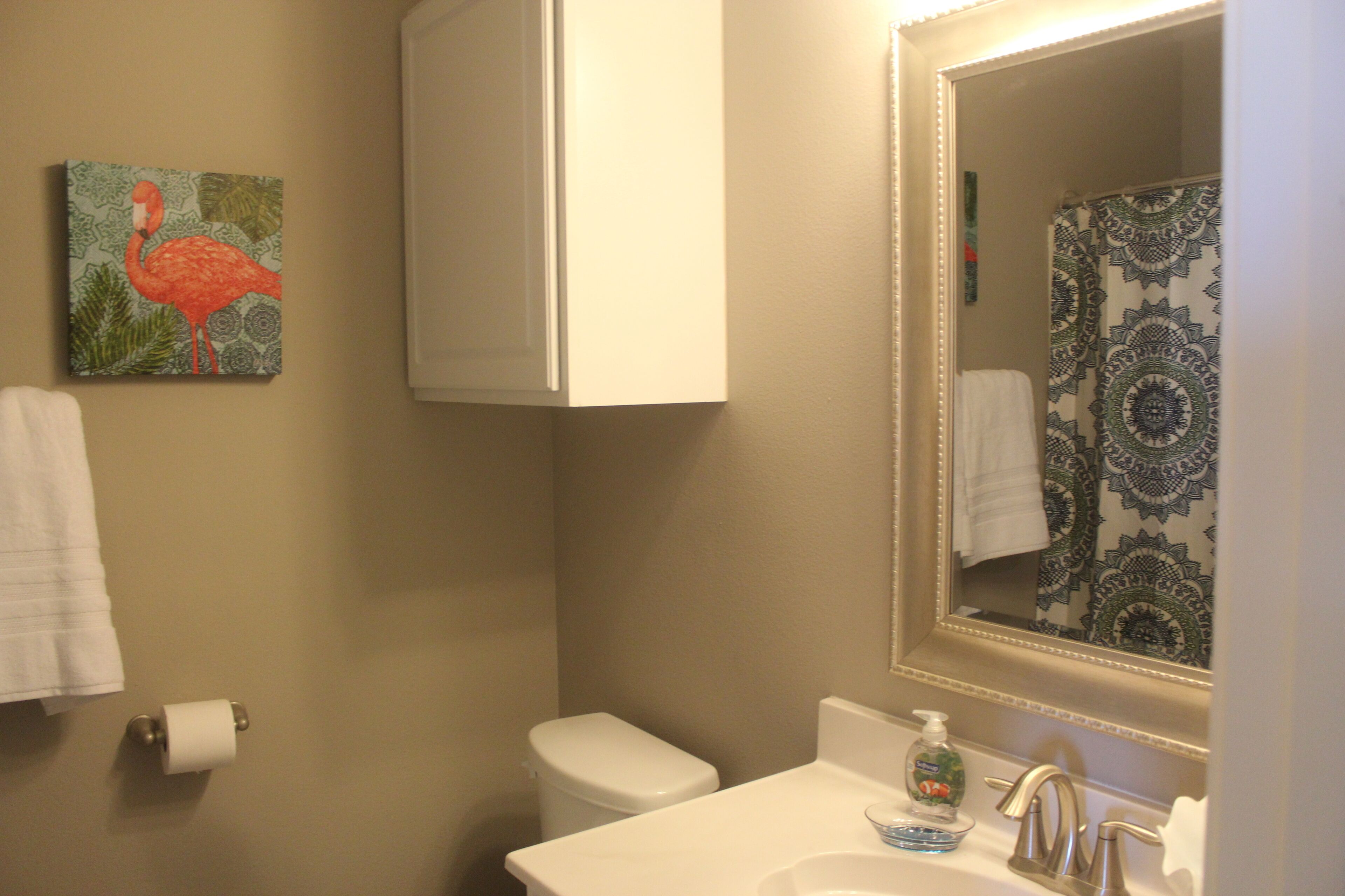 A bathroom with a toilet, sink, and mirror. There is a painting of a flamingo on the wall.