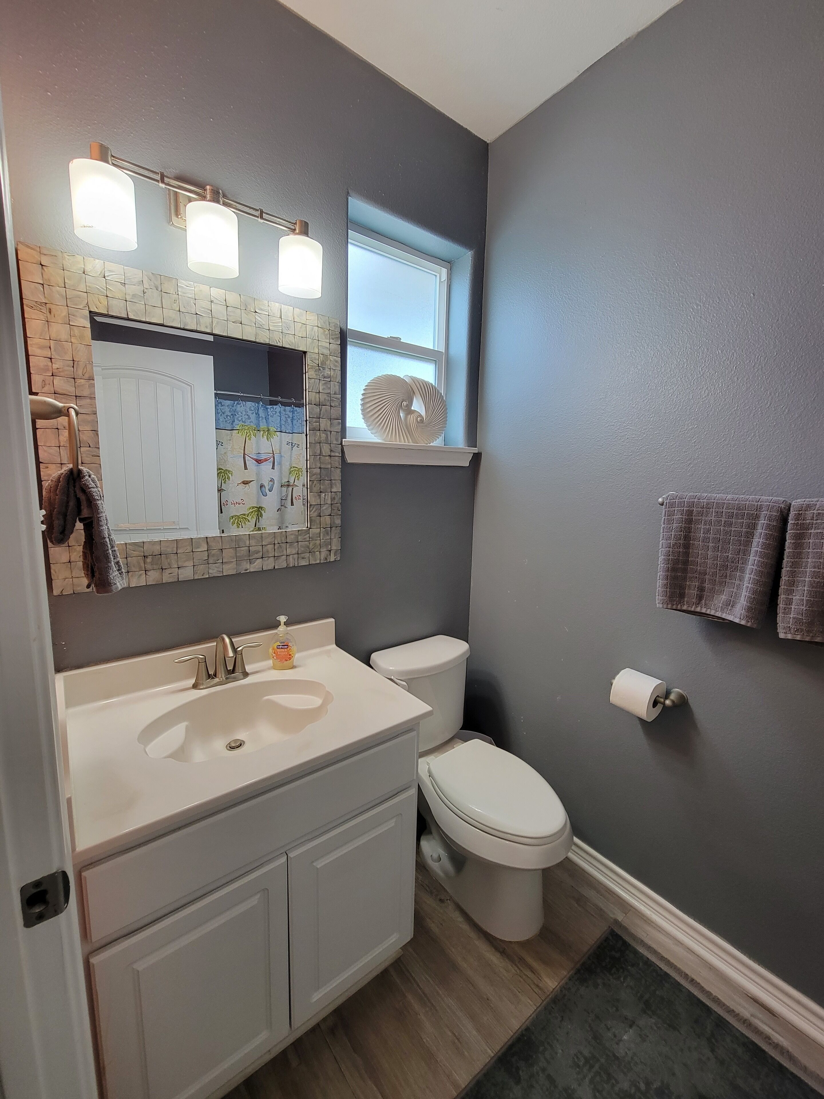 A bathroom with a white sink, a toilet, and a window with a decorative item. There are towels hanging on the wall and a shower curtain with a beach scene.