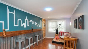 A room with a cityscape mural, a wooden bar counter with stools, a microphone stand, and a dining table with chairs.