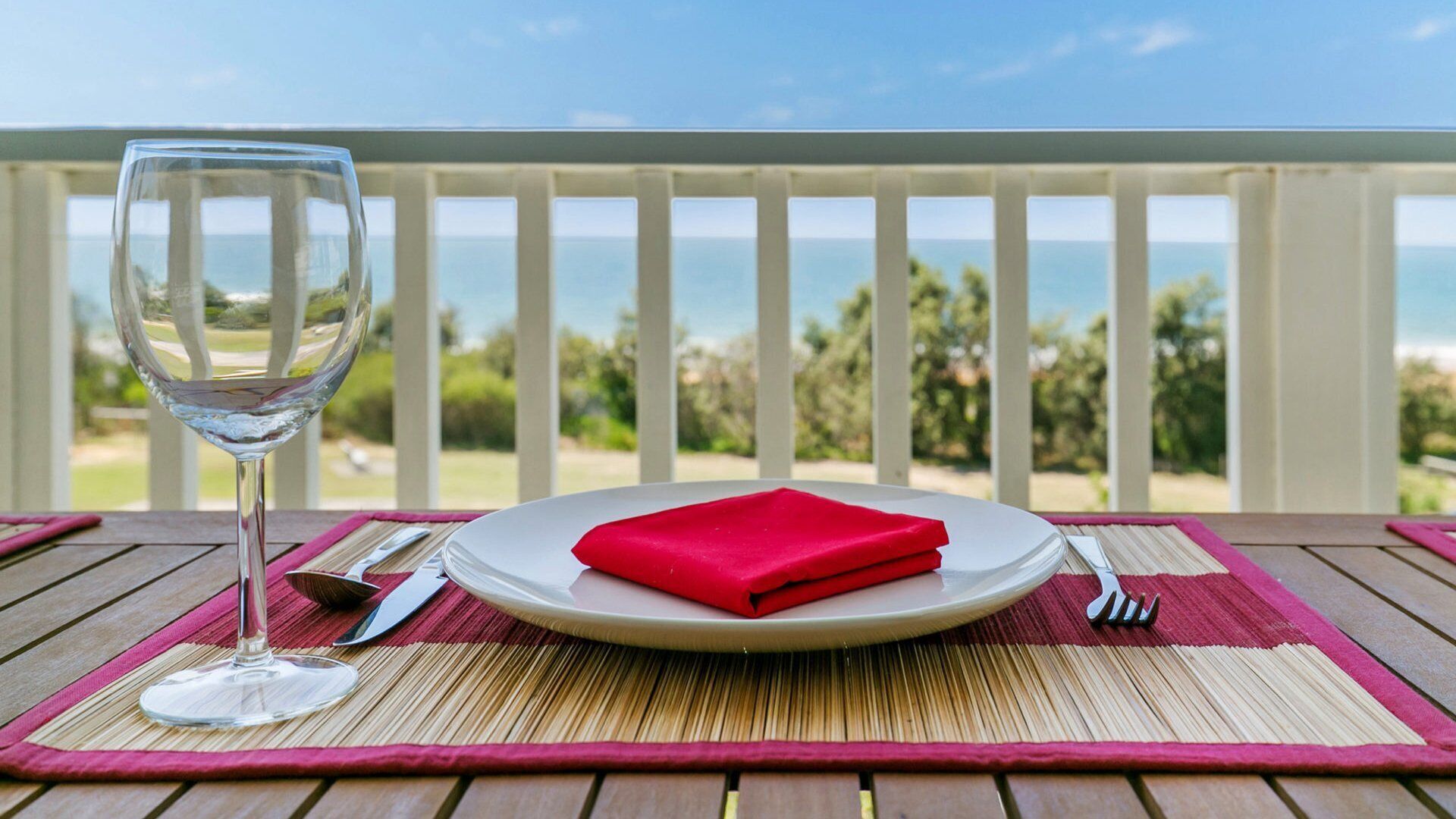 A table with a glass of white wine, a folded red napkin, and cutlery on a wooden table with a view of the ocean.
