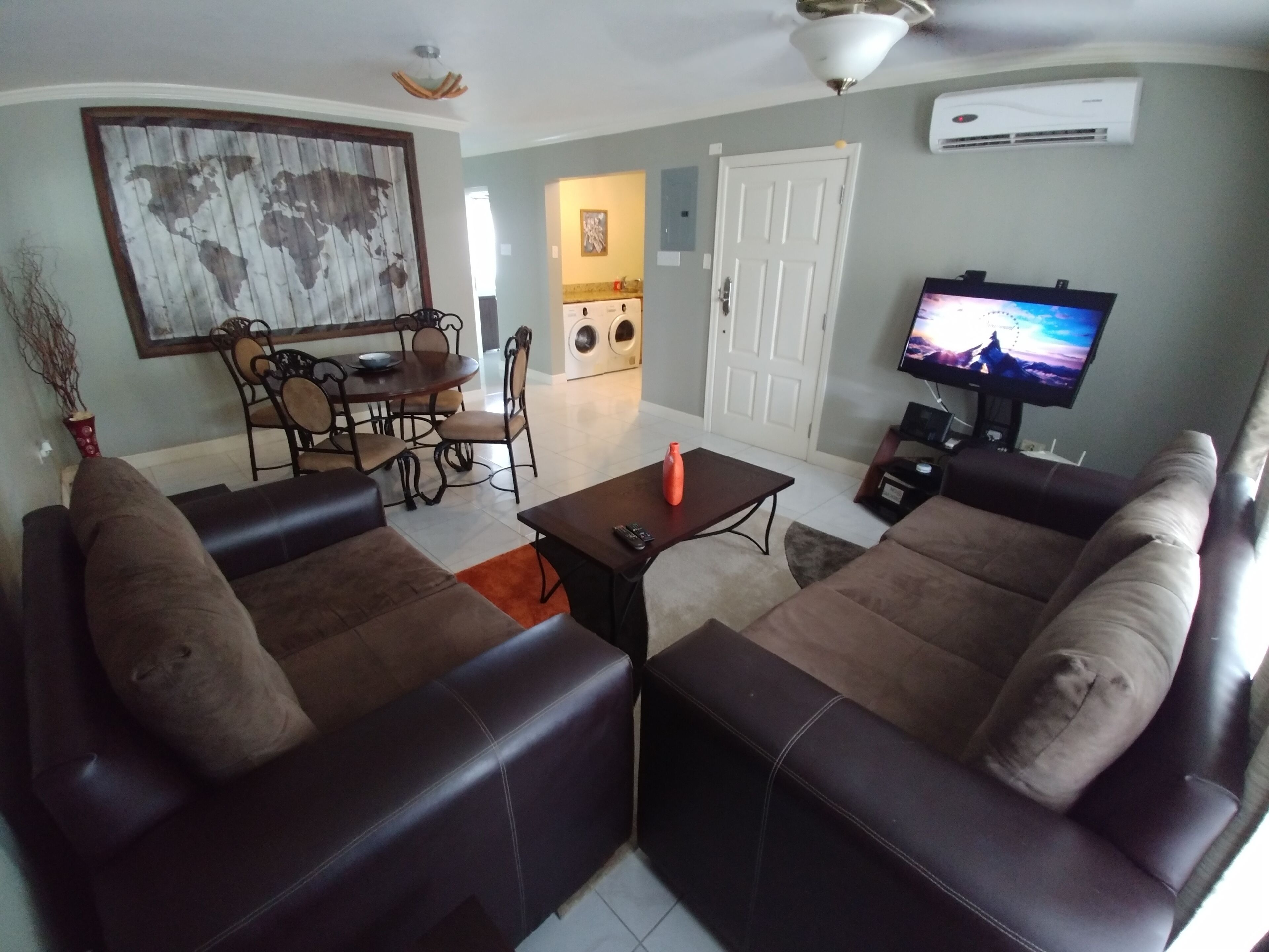 A living room with two brown sofas, a coffee table, a dining table with chairs, a TV, and an air conditioning unit.