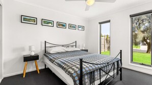 A bedroom with a bed, two bedside tables, a ceiling fan, and a large window with a view of a backyard. - Casa Killara - sleeps 14 (COWES)