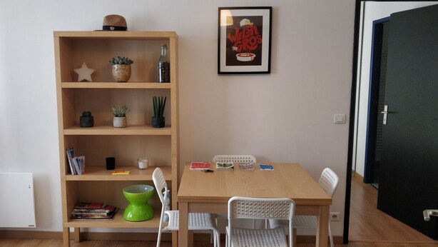 Une pièce avec une bibliothèque en bois, une affiche encadrée, une table avec des chaises et une porte.