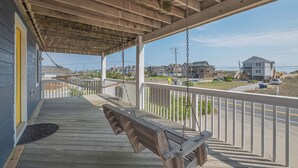 Una terraza de madera con un columpio, baranda blanca y vistas a casas y una carretera al fondo.