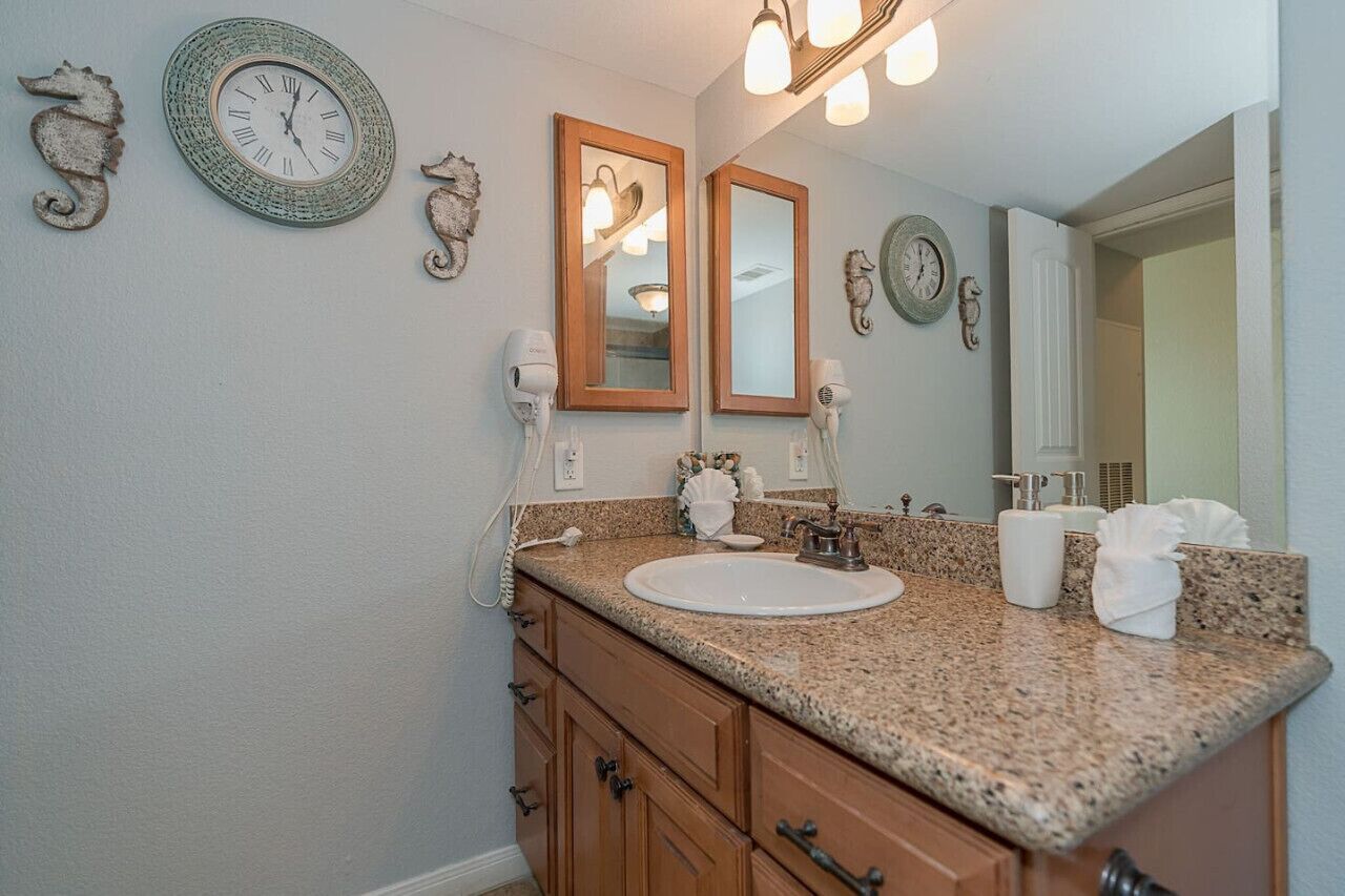 Un baño con encimera de granito, un lavabo, dos espejos y relojes decorativos en la pared.
