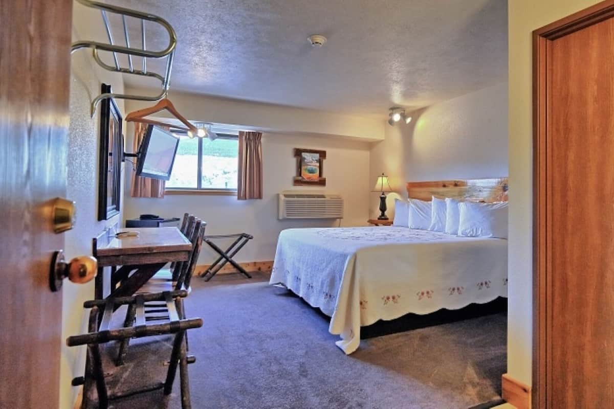 A bedroom with a bed, a desk, and a chair. There is a window with curtains, a wall-mounted heater, and a picture on the wall.
