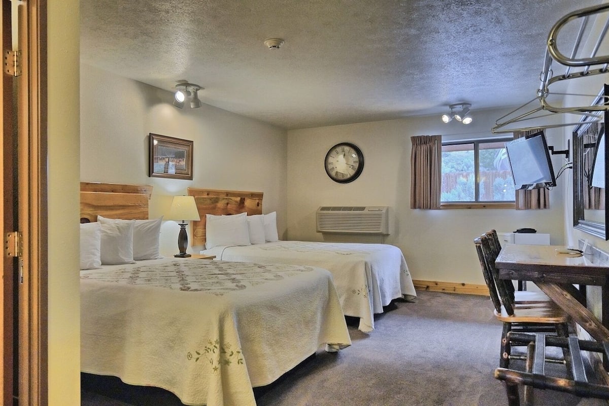 A hotel room with two beds, a desk, a chair, a clock, and a window.