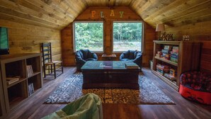 A cozy cabin interior with a wooden ceiling, two windows offering a view of the forest, a play area with a table and chairs, and a bookshelf filled with board games.