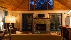 A cozy living room with a stone fireplace, wooden ceiling, and a large window overlooking trees.