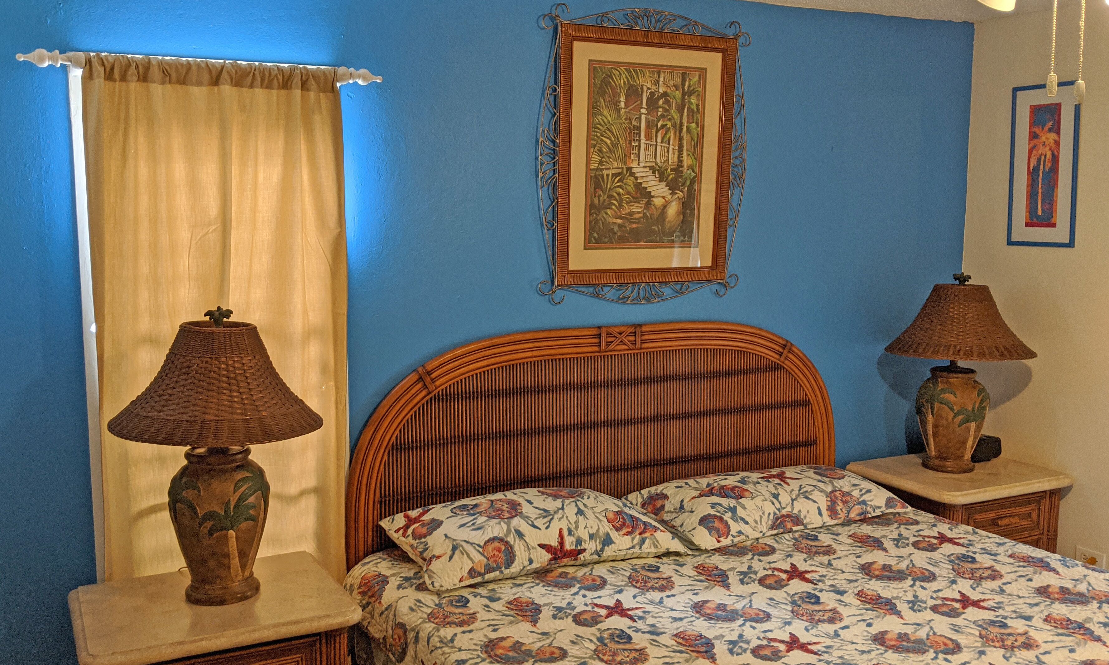 A bedroom with a bed, two nightstands, a window with curtains, a framed picture on the wall, and a lamp on each nightstand.