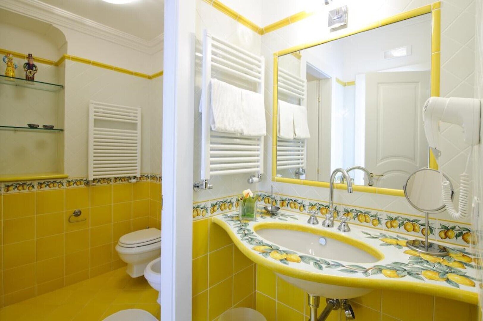 Une salle de bain avec des carreaux jaunes, un lavabo blanc orné d’un motif citron, un miroir et des toilettes blanches.