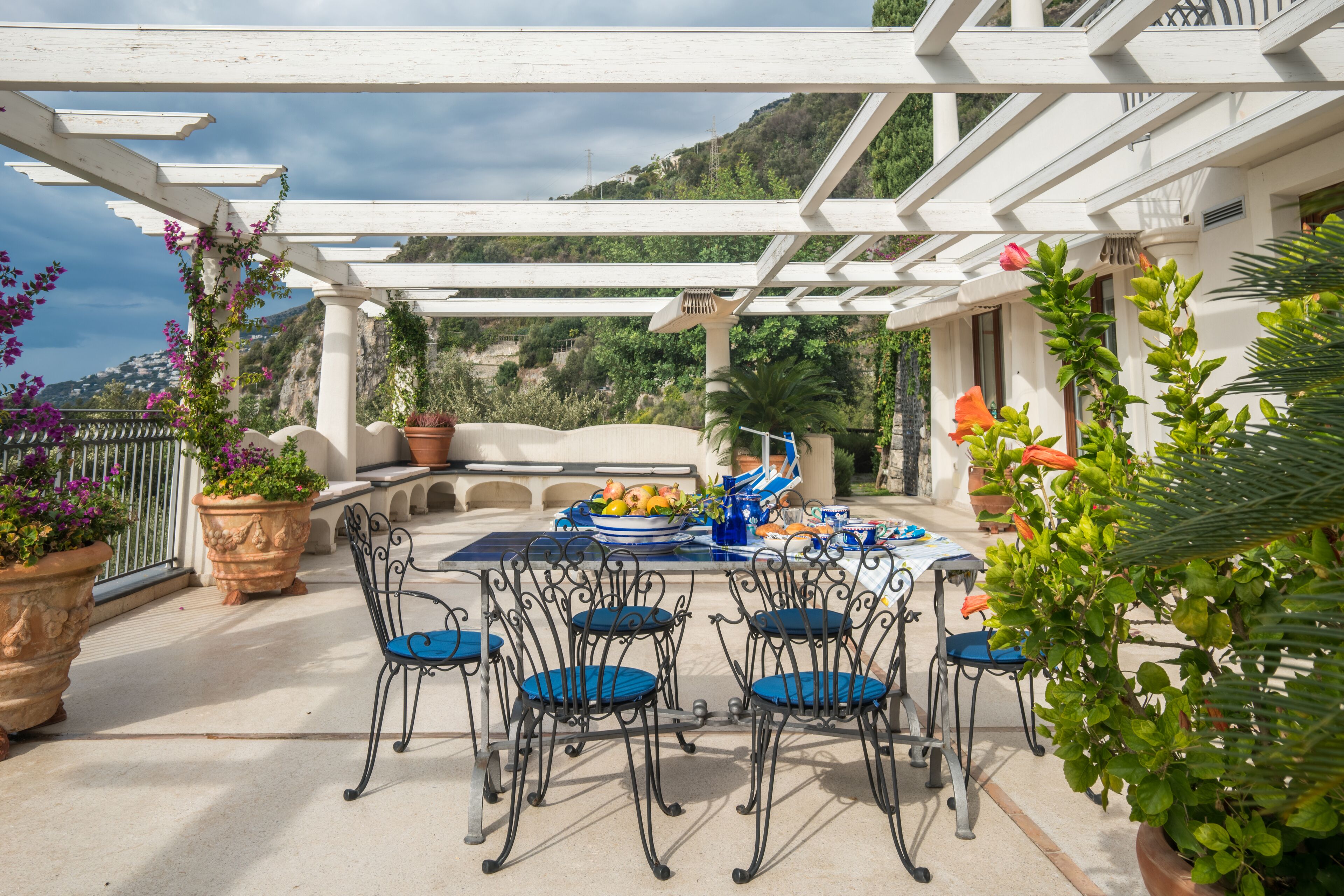 Een terras buiten met een witte pergola, een eettafel en potplanten.