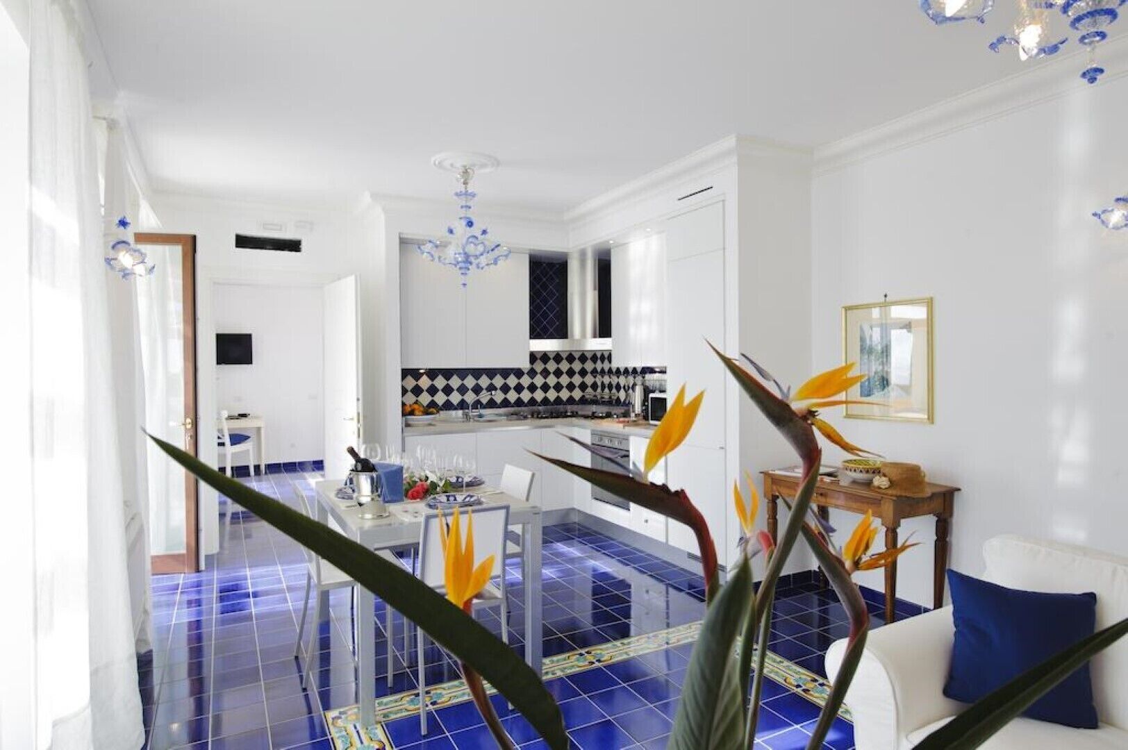 A modern kitchen with a black and white checkered backsplash, a dining area with a glass table, and a living room with a white sofa and blue cushions.