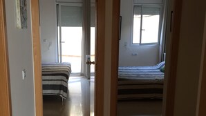 Ein Flur, der zu zwei Schlafzimmern mit gestreifter Bettwäsche führt. Ein Fenster mit Jalousien und eine Tür mit Messingklinke sind ebenfalls zu sehen.