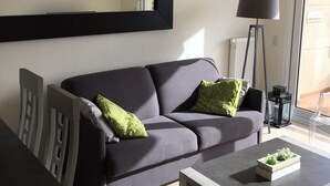 Ein modernes Wohnzimmer mit einem grauen Sofa, zwei grünen Kissen, einer schwarzen Stehlampe und einem Couchtisch mit einer dekorativen Schachtel.