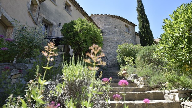 A stone building with a staircase leading to a door, surrounded by lush greenery and flowering plants. - Mazet de La Tour - private terrace and shared swimming pool -Vallée des Baux (Paradou)