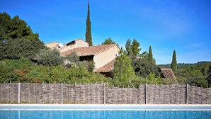 A swimming pool with a wooden fence in the foreground, a building with a tiled roof, and tall trees in the background. - Mazet de La Tour - private terrace and shared swimming pool -Vallée des Baux (Paradou)