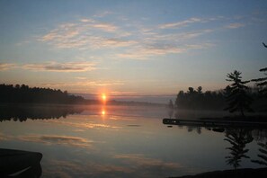 A calm lake at sunrise with the sun reflecting on the water, surrounded by trees and a misty atmosphere. - The Loon Muskoka Cabin - TeaLakeCottages (Coldwater)