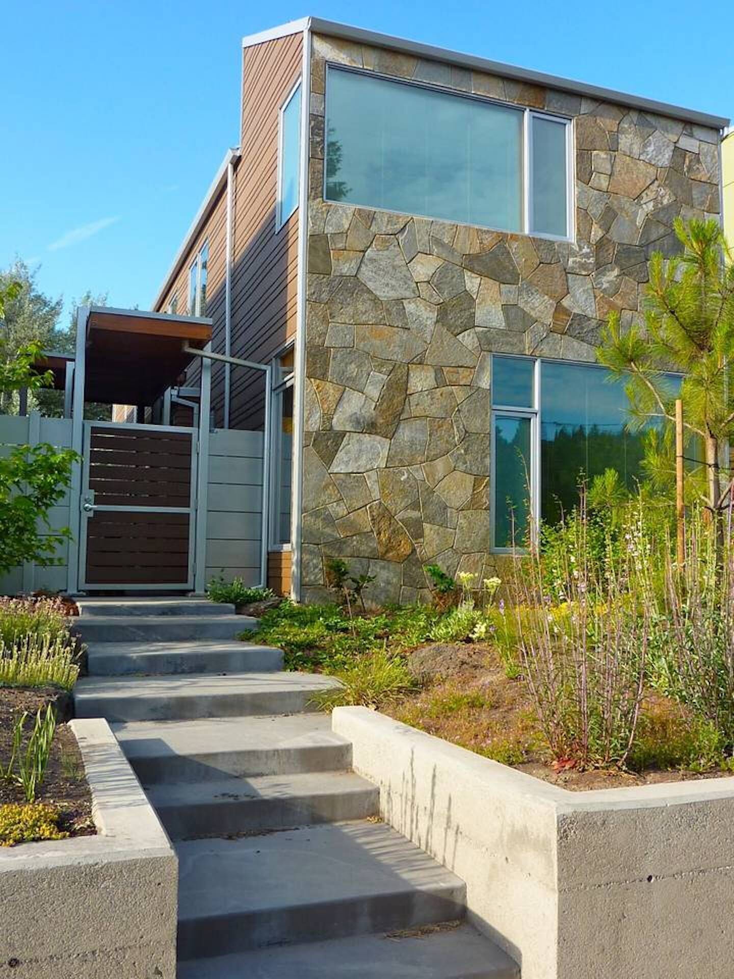 A modern house with a stone facade, a wooden deck, and a concrete staircase leading to a gated entrance.